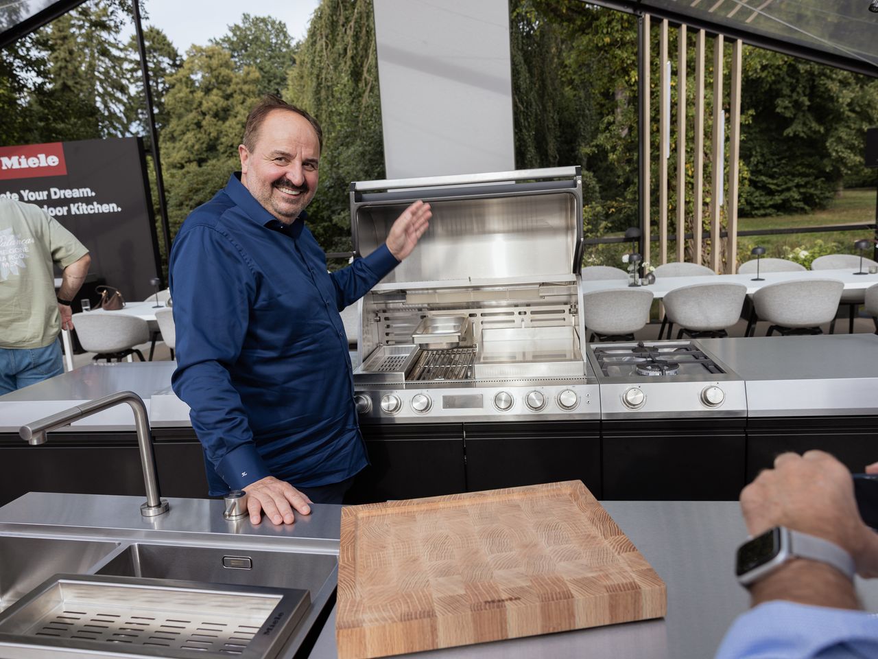 Spitzenkoch Johann Lafer steht l&auml;chelnd an einer ge&ouml;ffneten Edelstahl-Outdoor-K&uuml;che von Miele und zeigt begeistert auf die Ausstattung.