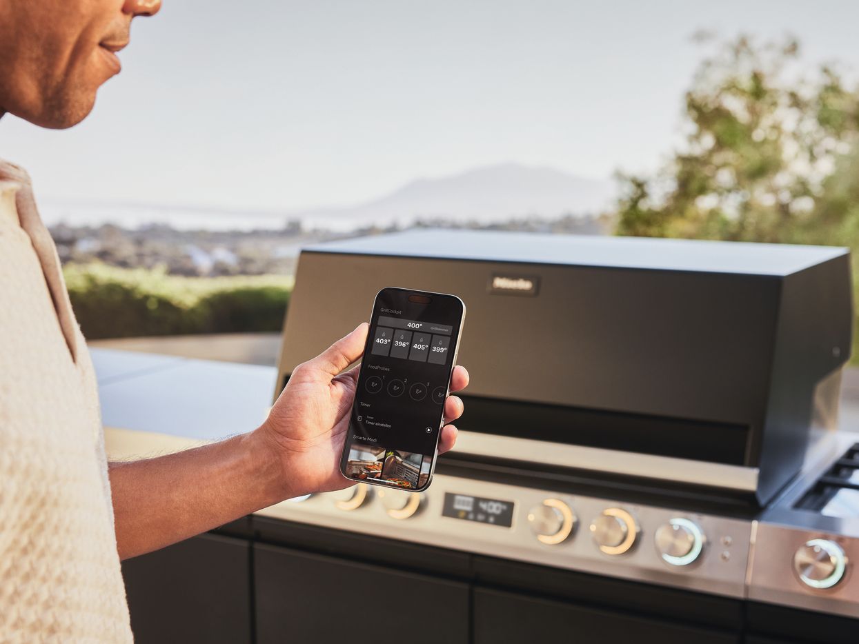 Nahaufnahme einer Hand, die ein Smartphone vor einer modernen Miele Outdoor-K&uuml;che h&auml;lt. Auf dem Display der App sind die genauen Temperaturzonen und Grilleinstellungen zu sehen.