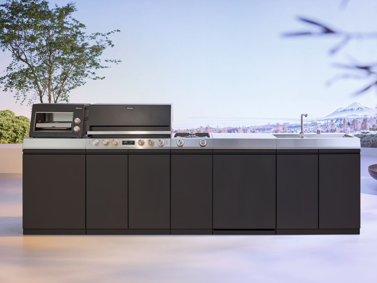 Die komplette Miele Outdoor-K&uuml;che &bdquo;Dreams&ldquo; in Anthrazit und Edelstahl, freigestellt vor einer hellen, sommerlichen Horizont-Kulisse.
