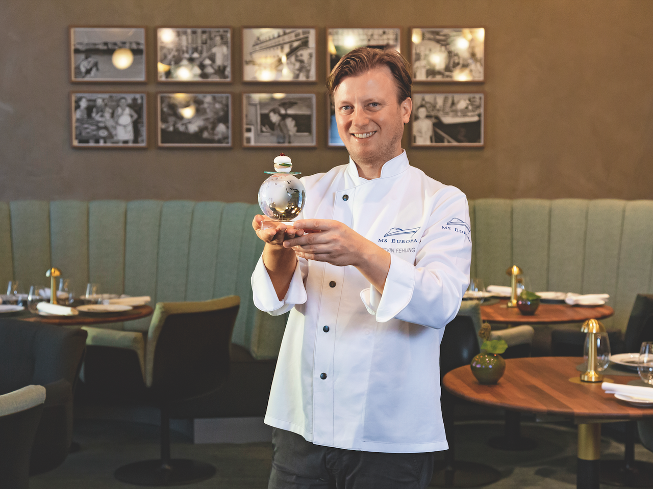 Kevin Fehling steht im Restaurant THE GLOBE an Bord der EUROPA und h&auml;lt eine gl&auml;serne Weltkugel mit seinem Fischbr&ouml;tchen in den H&auml;nden.