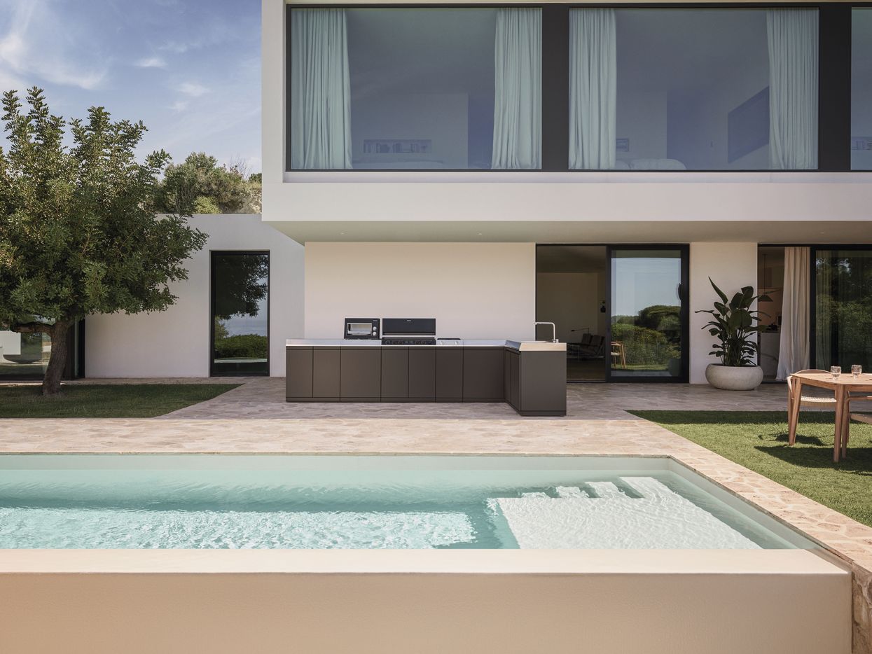 Eine minimalistische, moderne Outdoor-Küche von Miele steht auf einer Naturstein-Terrasse neben einem luxuriösen Pool vor einer Design-Villa im Sommer.