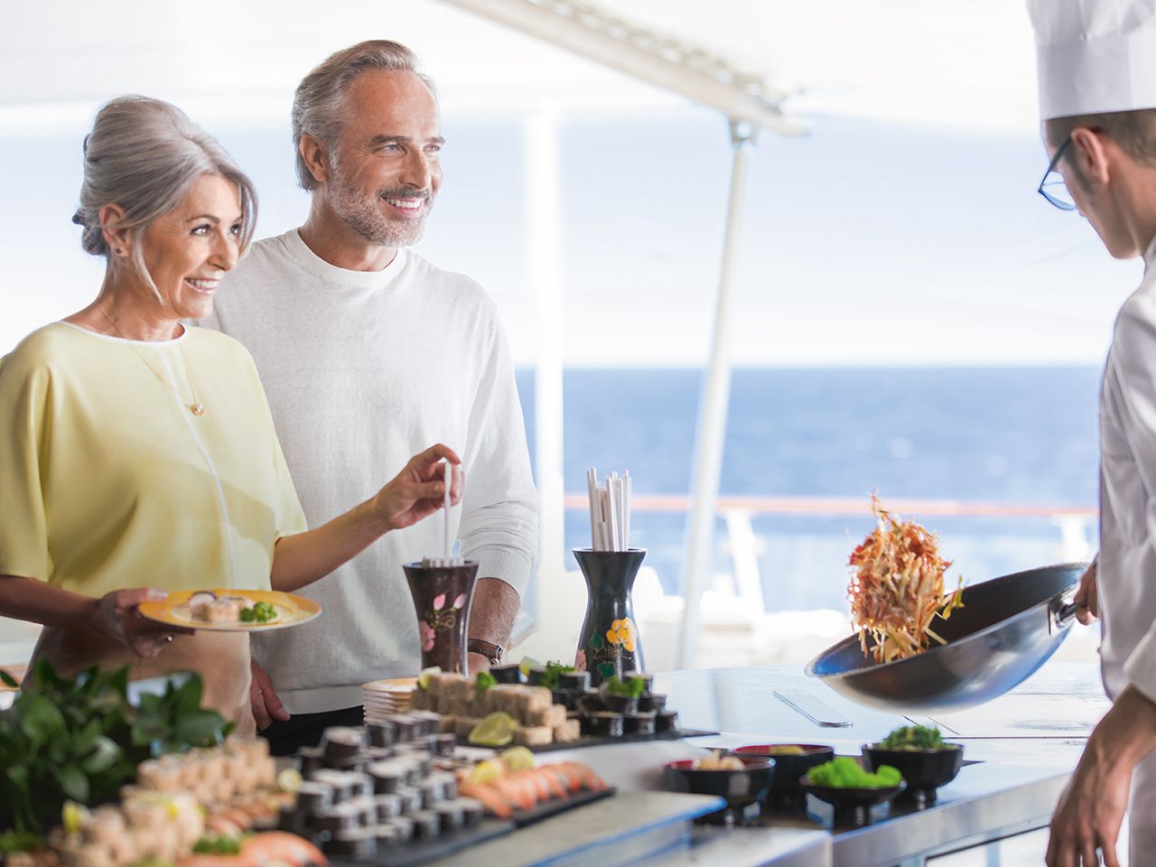G&auml;stepaar genie&szlig;t an Bord eines Hapag-Lloyd-Kreuzfahrtschiffs Sushi und weitere Speisen, w&auml;hrend ein Koch ein Gericht zubereitet. Im Hintergrund ist das Meer zu sehen.