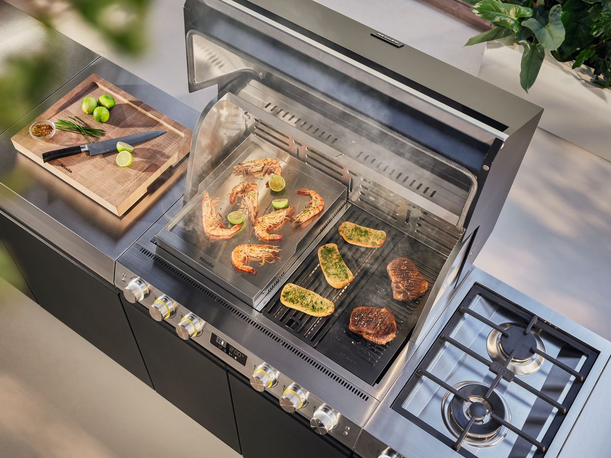 Nahaufnahme eines geöffneten, großen Edelstahl-Gasgrills, auf dessen Rost zeitgleich Riesengarnelen, Grillwürstchen, ganze Maiskolben und mit Speck umwickelter grüner Spargel brutzeln.
