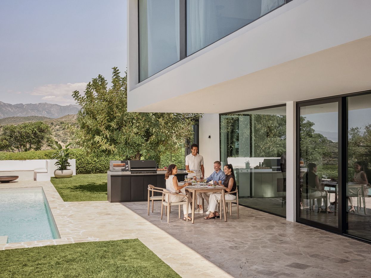 Eine kleine G&auml;sterunde sitzt lachend an einem stilvollen Holztisch auf einer sonnigen Terrasse am Pool, w&auml;hrend im Hintergrund an einer Outdoor-K&uuml;che gegrillt wird.